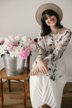Metal kova ile peonies rustik ahşap sandalye üzerinde oturan şapkalı şık boho kız. Büyük peonies buket gülümseyen güzel hipster kadın. Kırsal yaşam. Uluslararası Kadınlar günü