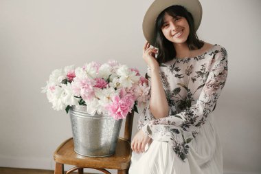 Metal kova ile peonies rustik ahşap sandalye üzerinde oturan şapkalı şık boho kız. Büyük peonies buket gülümseyen güzel hipster kadın. Kırsal yaşam. Uluslararası Kadınlar günü