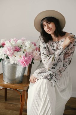 Büyük peonies buket ile poz şapkalı güzel esmer boho kız. Metal kova ile peonies rustik ahşap sandalye üzerinde oturan şık hipster kadın. Uluslararası Kadınlar günü