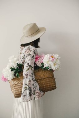 Arkadan görünüşü boho kız pembe ve beyaz peonies rustik sepet içinde tutarak. Şapka ve çiçeklerle poz bohem çiçek elbise şık hipster kadın. Uluslararası Kadınlar günü. Kırsal yaşam