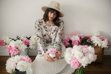 Pembe ve beyaz peonies rustik ahşap katta oturan boho kız portresi. Pembe şakayık çiçekleri arasında tutarak bohem içinde şık hipster kadın. Uluslararası Kadınlar günü