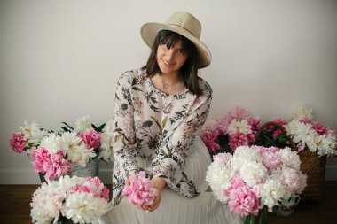 Pembe ve beyaz peonies rustik ahşap katta oturan boho kız portresi. Pembe şakayık çiçekleri arasında tutarak bohem içinde şık hipster kadın. Uluslararası Kadınlar günü