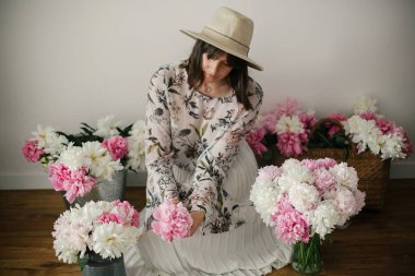 Pembe ve beyaz peonies rustik sepeti ve ahşap zemin üzerinde metal kova içinde oturan Boho kız. Çiçekler içinde bohem şık hipster kadın. Mutlu anneler günü.