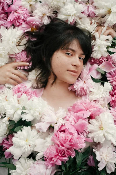 Güzel esmer kız çok pembe ve beyaz peonies yalan. Boho kadın portre şakayık çiçekleri doğal makyaj ile. Yaratıcı çiçek fotoğraf. Aroma kokusu kavramı. Uluslararası Kadınlar günü
