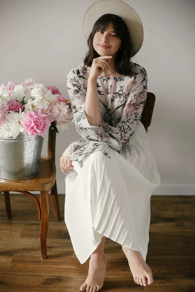 Büyük peonies buket ile poz şapkalı güzel esmer boho kız. Metal kova ile peonies rustik ahşap sandalye üzerinde oturan şık hipster kadın. Uluslararası Kadınlar günü