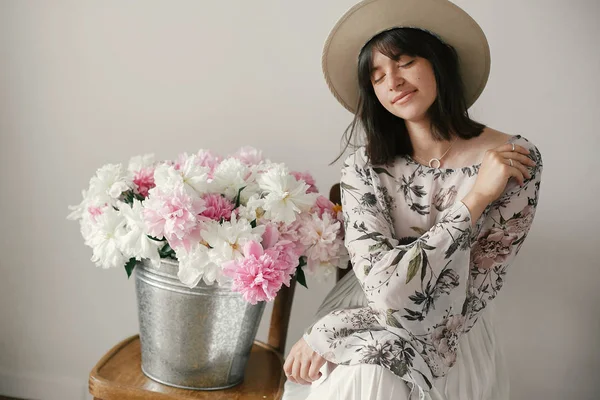 Metal kova ile peonies rustik ahşap sandalye üzerinde oturan şapkalı şık boho kız. Güzel hipster kadın büyük peonies buket ile poz. Kırsal yaşam. Uluslararası Kadınlar günü