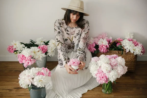 Pembe ve beyaz peonies rustik sepeti ve ahşap zemin üzerinde metal kova içinde oturan Boho kız. Pembe şakayık çiçekleri arasında tutarak bohem içinde şık hipster kadın. Kırsal yaşam