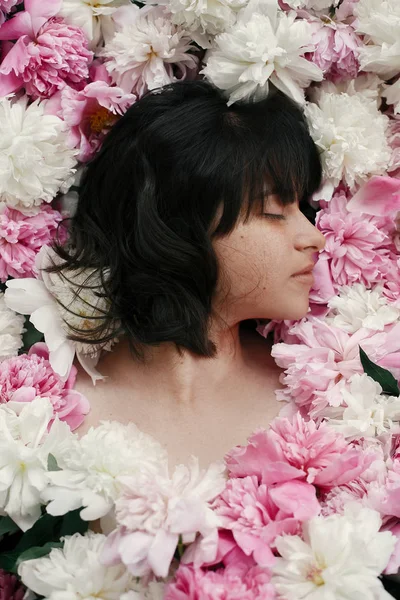 Güzel esmer kız çok pembe ve beyaz peonies yalan. Boho kadın portre şakayık çiçekleri doğal makyaj ile. Yaratıcı çiçek fotoğraf. Aroma kokusu kavramı. Uluslararası Kadınlar günü
