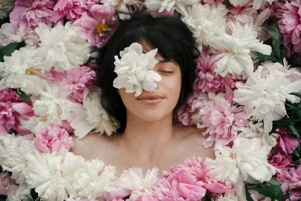 Beautiful brunette girl with white peony on eye lying in pink peonies. Aroma and care concept. International Womens Day. Creative art floral photo. Portrait of boho woman in many flowers