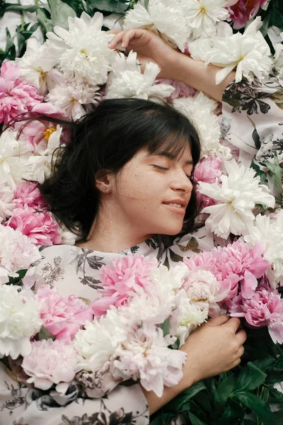 Portrait of boho woman with natural makeup lying in peonies. Creative floral photo. Aroma and spa concept. International Womens Day. Beautiful brunette girl in many pink and white flowers
