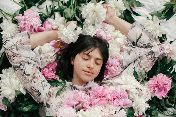 Portrait of boho woman with natural makeup lying in peonies. Creative floral photo. Aroma and spa concept. International Womens Day. Beautiful brunette girl in many pink and white flowers