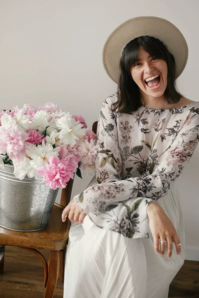 Oturma ve metal kova peonies rustik ahşap sandalye ile komik şapkalı şık boho kız. Güzel hipster kadın büyük peonies buket ile poz. Kırsal yaşam.
