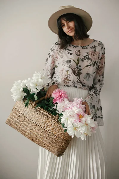 Pembe ve beyaz peonies rustik sepet içinde tutarak şapkalı Boho kız. Çiçeklerle poz bohem Çiçek elbiseli şık hipster kadın. Uluslararası Kadınlar günü. Kırsal yaşam