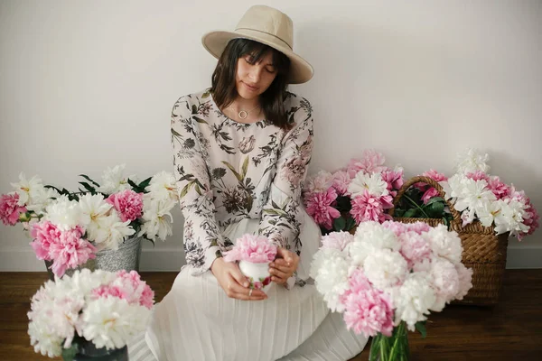 Pembe ve beyaz peonies rustik ahşap katta oturan boho kız portresi. Pembe şakayık çiçekleri arasında tutarak bohem içinde şık hipster kadın. Uluslararası Kadınlar günü