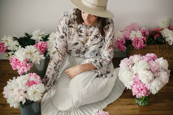 Pembe ve beyaz peonies rustik sepeti ve ahşap zemin üzerinde metal kova içinde oturan Boho kız. Çiçekler içinde bohem şık hipster kadın. Mutlu anneler günü.