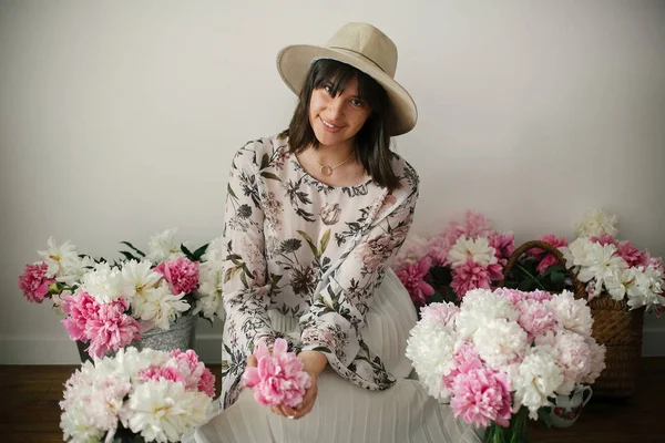 Pembe ve beyaz peonies rustik ahşap katta oturan boho kız portresi. Pembe şakayık çiçekleri arasında tutarak bohem içinde şık hipster kadın. Uluslararası Kadınlar günü