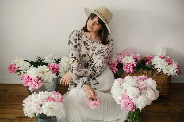 Pembe ve beyaz peonies rustik sepeti ve ahşap zemin üzerinde metal kova içinde oturan Boho kız. Pembe şakayık çiçekleri arasında tutarak bohem içinde şık hipster kadın. Kırsal yaşam