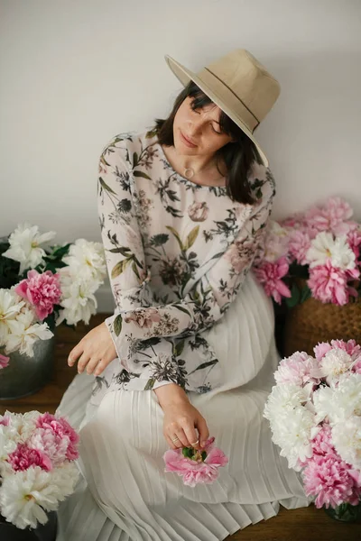 Pembe ve beyaz peonies rustik sepeti ve ahşap zemin üzerinde metal kova içinde oturan Boho kız. Çiçekler içinde bohem şık hipster kadın. Mutlu anneler günü.