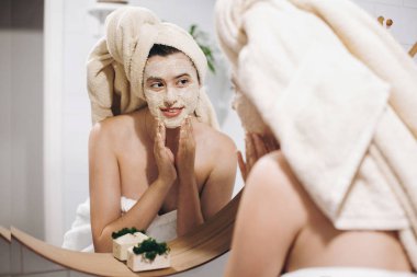 Organik yüz maskesi uygulamak ve şık banyo yuvarlak aynaya bakarak havlu genç mutlu kadın. Soyma ve yüzünde cilt temizliği ile fırçalayın, yüz masajı yapma kız. Cilt Bakımı