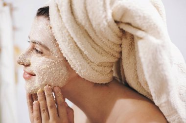 Skin Care concept. Young happy woman in towel making facial massage with organic face scrub close up in stylish bathroom. Girl applying scrub cream, peeling and cleaning skin. clipart