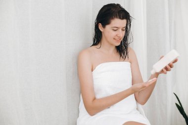 Beyaz havlu uygulanan nemlendirici içinde genç bir kadın olarak ev banyo yeşil bitkiler ile yandan krem. Cilt ve vücut bakımı. Losyon ile plastik şişe tutan el. Spa ve sağlıklı yaşam