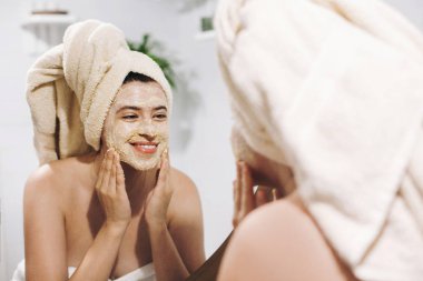 Skin Care concept. Young happy woman in towel making facial massage with organic face scrub and looking at mirror in stylish bathroom. Girl applying scrub cream, peeling and cleaning skin clipart