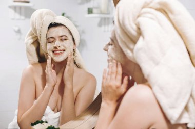 Organik yüz maskesi uygulamak ve şık banyo yuvarlak aynaya bakarak havlu genç mutlu kadın. Soyma ve yüzünde cilt temizliği ile fırçalayın, yüz masajı yapma kız. Cilt Bakımı