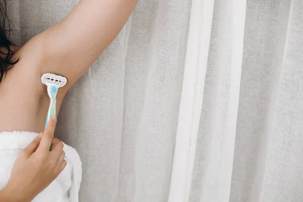 Hand holding plastic razor and shaving hairy armpit. Young woman shaving armpits with plastic razor closeup in home bathroom. Skin care. Hair Removal concept. Copy space