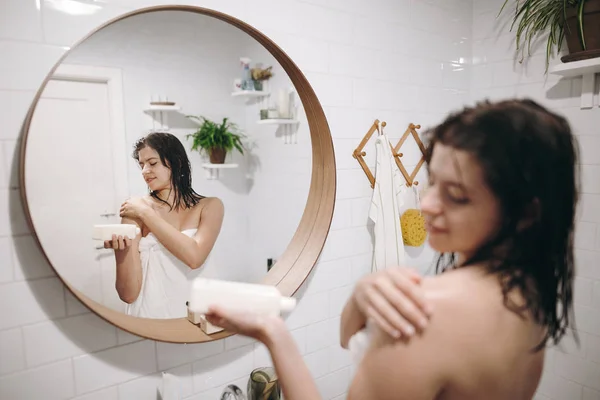 白いタオルを浴室で肩 ミラーで反射に保湿クリームを適用することで 若い幸せな女性 皮膚及び身体の手入れ ハンド ローションとプラスチック ボトル セクシーな女性のリラックス — ストック写真