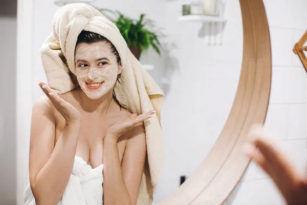 Young happy woman in towel applying organic face scrub and looking at round mirror in stylish bathroom. Girl making facial massage, peeling and cleaning skin on face. Skin Care and Hygiene