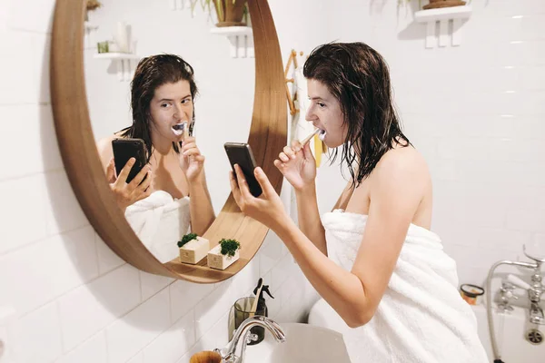 毎日の朝の日課 歯を磨くと丸い鏡でスタイリッシュな浴室での携帯電話の画面を見て白いタオルで若い幸せな女 自然な肌と濡れた髪でスリムなセクシーな女性 — ストック写真