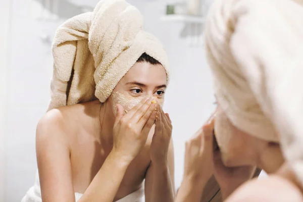 Organik yüz fırçalayın ile yüz masajı yapma ve şık banyo aynaya bakarak havlu genç mutlu kadın. Uygulama kız krem, peeling ve temizlik cilt fırçalayın. Cilt Bakımı