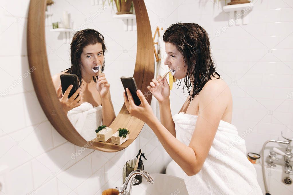 Daily morning routine. Young happy woman in white towel brushing teeth and looking at phone screen in stylish bathroom at round mirror. Slim sexy woman with natural skin and wet hair