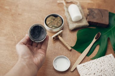 Sıfır atık. Doğal çevre kömür diş macunu cam bambu diş fırçası, sabun, katı şampuan, metal ustura, deodorant yeşil monstera yaprak ile ahşap arka plan üzerinde tutan el.