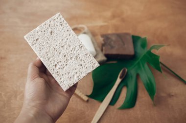 Doğal çevre sünger bambu diş fırçası, sabun, katı şampuan, kristal deodorant, yeşil monstera yaprak ile ahşap arka plan üzerinde tutan el. Sıfır atık kavramı. Sürdürülebilir yaşam tarzı