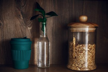 Sıfır atık kavramı, sürdürülebilir yaşam tarzı. Cam kavanoz granola, yeniden kullanılabilir kahve fincanı ve bambu yaprakları ahşap raf üzerinde toplu alışveriş mağazası. Yasak Oda plastik.