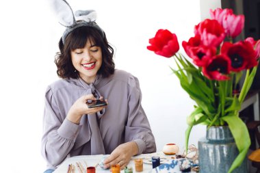 Tavşan kulakları Boyalı Paskalya yumurta masada lale vazo ve gülümseyen ile telefonda fotoğrafta güzel kız. Paskalya yumurtası dekorasyon şık genç esmer kadın. Mutlu Paskalya