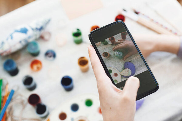 Hands holding phone and taking photo of stylish easter flat lay of painting egg on rustic table with paint,brushes, bird toy. Easter workshop.Decorating easter egg, holiday preparations.