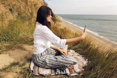 Pratik yoga plajda, otların arasında oturan ve seyir telefon genç güzel kadın selfie alarak. Hippi kız yoga, rahatlatıcı ve meditatiig yapıyor. Uluslararası Yoga gün kavramı.