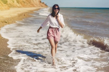 Yaz tatil kavramı. Beyaz gömlek ve çalıştıran ve dalgalar ile eğleniyor güneşli kumsalda güneş gözlüğü mutlu genç kadın. Rahatlatıcı ve denizde gülümseyen hipster ince kız. Uzay kopyalayın. Neşeli an