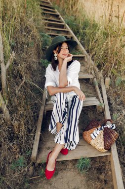 Şık şık görünüm denizde güneşli ışıkta beach cliff ahşap merdivenlerde oturan kız. Mutlu genç boho kadın hasır çanta batımında sahilde rahatlatıcı ile şapkalı. Yaz tatili.