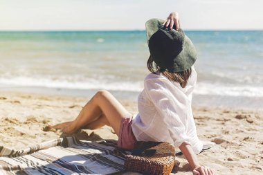 Yaz tatil kavramı. Mutlu genç kadın rahatlatıcı sahilde. Hipster ince kız beyaz gömlek ve oturma ve deniz kenarında sahilde dalgalar, güneşli sıcak hava ile bronzlaşma şapka. Huzurlu sakin an