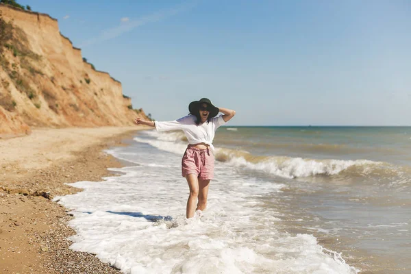 Beyaz gömlek ve çalıştıran ve dalgalar ile eğleniyor güneşli kumsalda şapka mutlu genç kadın. Rahatlatıcı ve denizde gülümseyen hipster ince kız. Yaz tatil kavramı. Uzay kopyalayın. Neşeli an