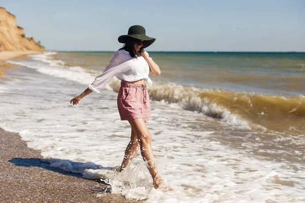 Yaz tatil kavramı. Beyaz gömlek ve çalıştıran ve dalgalar ile eğleniyor güneşli kumsalda şapka mutlu genç kadın. Rahatlatıcı ve denizde gülümseyen hipster ince kız. Uzay kopyalayın. Neşeli an