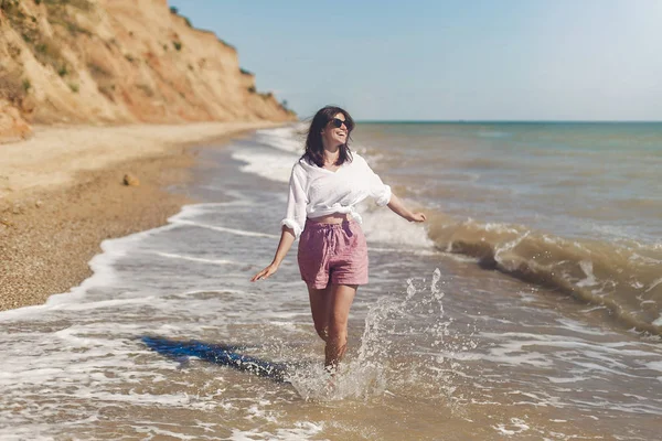 Yaz tatil kavramı. Beyaz gömlek ve çalıştıran ve dalgalar ile eğleniyor güneşli kumsalda güneş gözlüğü mutlu genç kadın. Rahatlatıcı ve denizde gülümseyen hipster ince kız. Uzay kopyalayın. Neşeli an