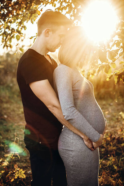 Stylish pregnant couple holding hands on belly and embracing in sunny light in autumn park under tree. Happy young parents, mom and dad, hugging baby bump, enjoying beautiful moment at sunset