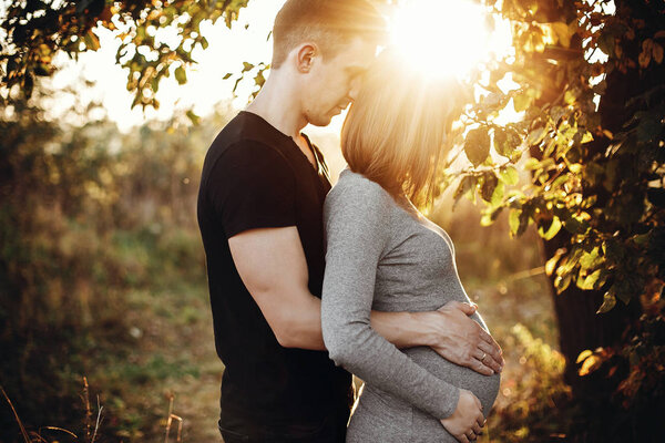 Stylish pregnant couple holding hands on belly in sunny light in autumn park. Happy young parents, mom and dad, hugging baby bump, smiling, enjoying beautiful moment at sunset. Parenthood concept