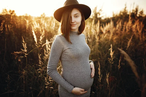 Stylish pregnant woman holding hands on belly in sunny light in — Stock Photo, Image
