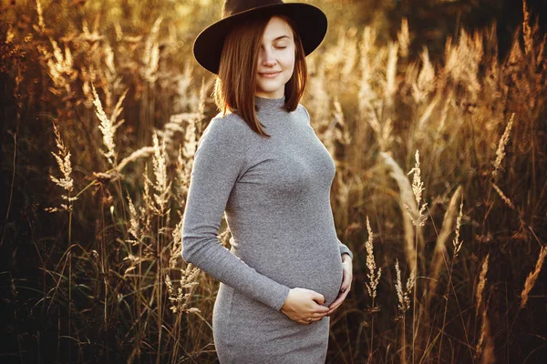 Stylish pregnant woman holding hands on belly in sunny light in — Stock Photo, Image