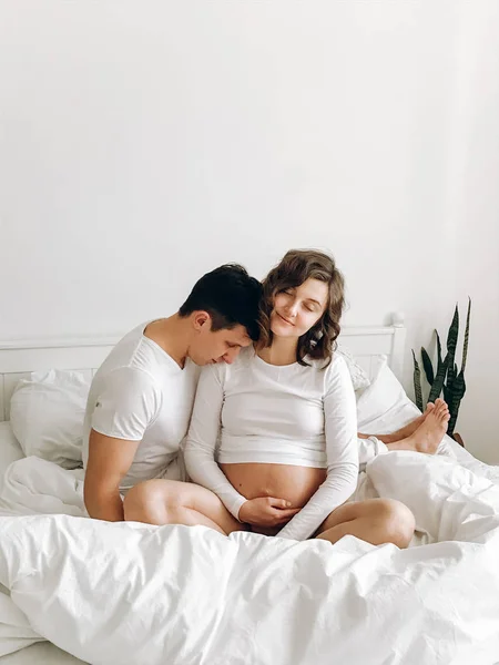 Happy νεαρό ζευγάρι έγκυος κρατώντας την κοιλιά χτύπημα σε λευκό κρεβάτι. STY — Φωτογραφία Αρχείου